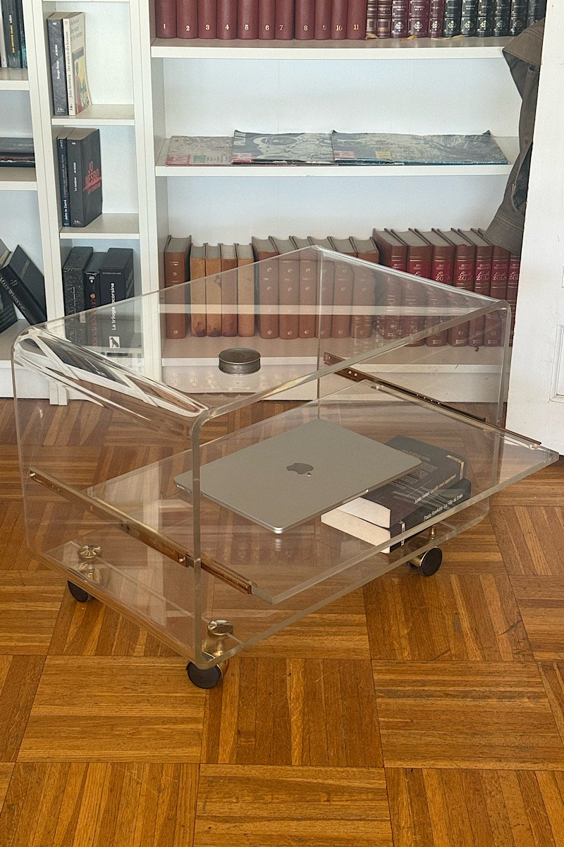 Vue de profil d'une desserte en plexiglas transparent, montrant la structure latérale, les roulettes et les plateaux sur un sol en parquet