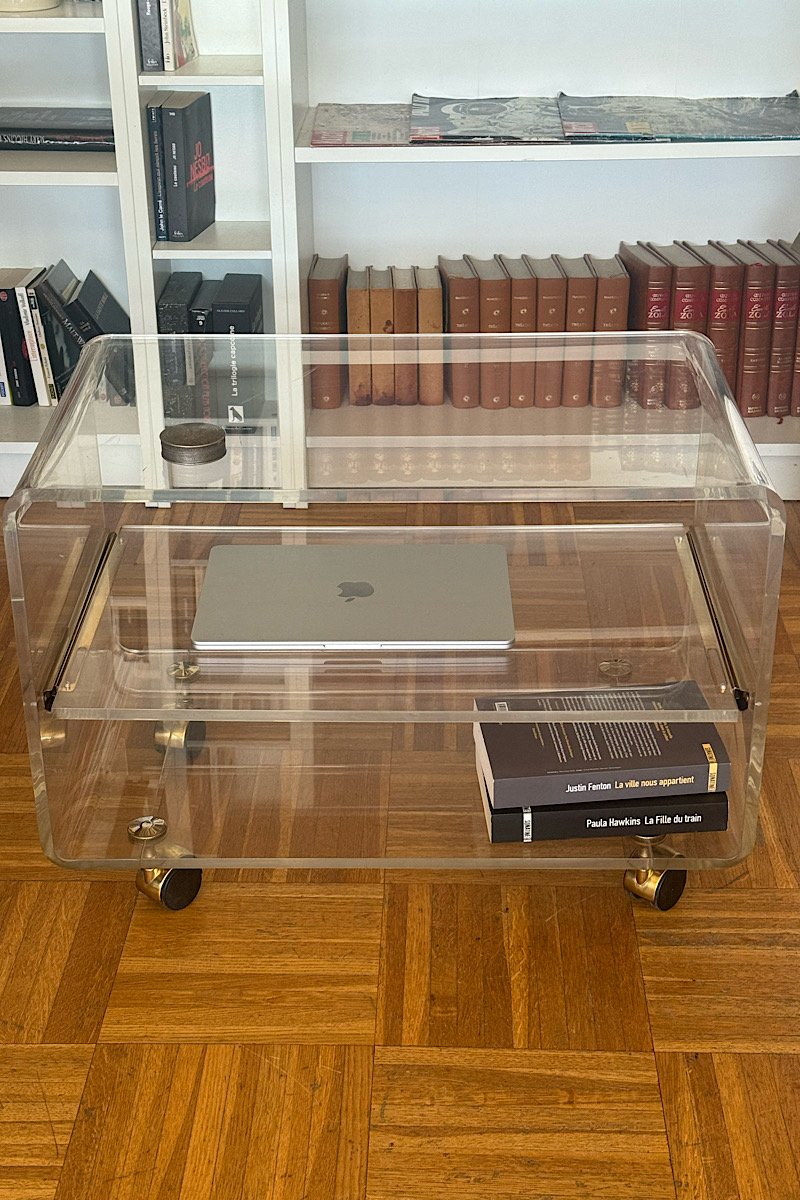 Vue de dessus d'une desserte en plexiglas transparent laissant apparaître le parquet en bois en dessous, avec une bibliothèque en arrière-plan