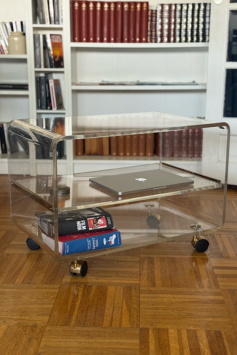 Une desserte vintage avec une tablette coulissante intermédiaire en plexiglas et métal sur roulettes, posée sur un parquet en bois devant une bibliothèque blanche