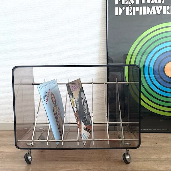 Un range-vinyles en plexiglas sur roulettes garni de quelques disques 33 tours, posé sur un parquet en bois