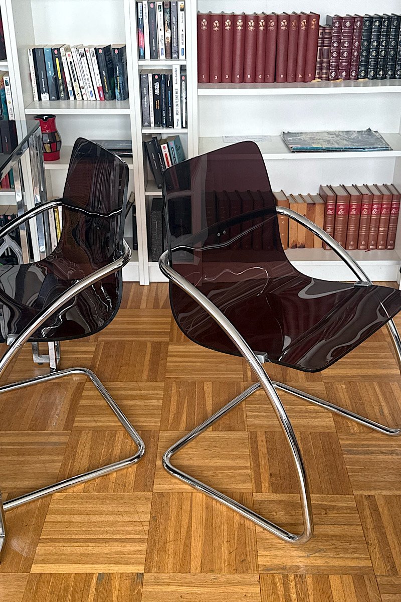 Deux chaises design "Blooming" créées par Yves Christin, mettant en évidence leur structure métallique tubulaire et leur assise en plexiglas fumé dans un intérieur avec bibliothèque.