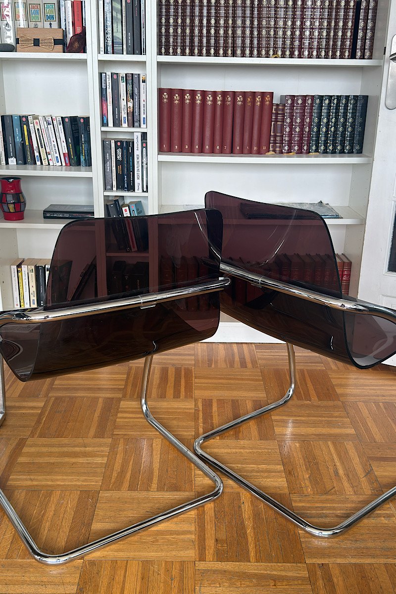 Vue de dos de deux chaises design "Blooming" créées par Yves Christin, mettant en évidence leur structure métallique tubulaire et leur assise en plexiglas fumé dans un intérieur avec bibliothèque