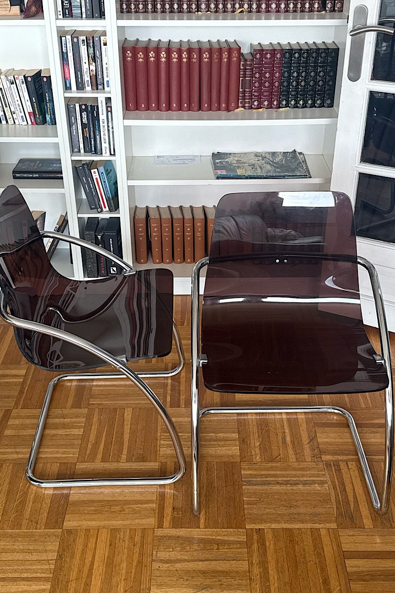 Deux chaises design "Blooming" créées par Yves Christin, présentées dans un intérieur avec parquet, soulignant le contraste entre la structure métallique tubulaire et l'assise en plexiglas fumé.