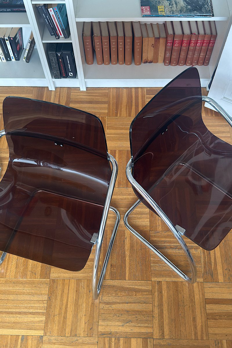Vue de dessus de deux chaises design "Blooming" créées par Yves Christin, mettant en évidence leur structure métallique tubulaire et leur assise en plexiglas fumé dans un intérieur avec bibliothèque.