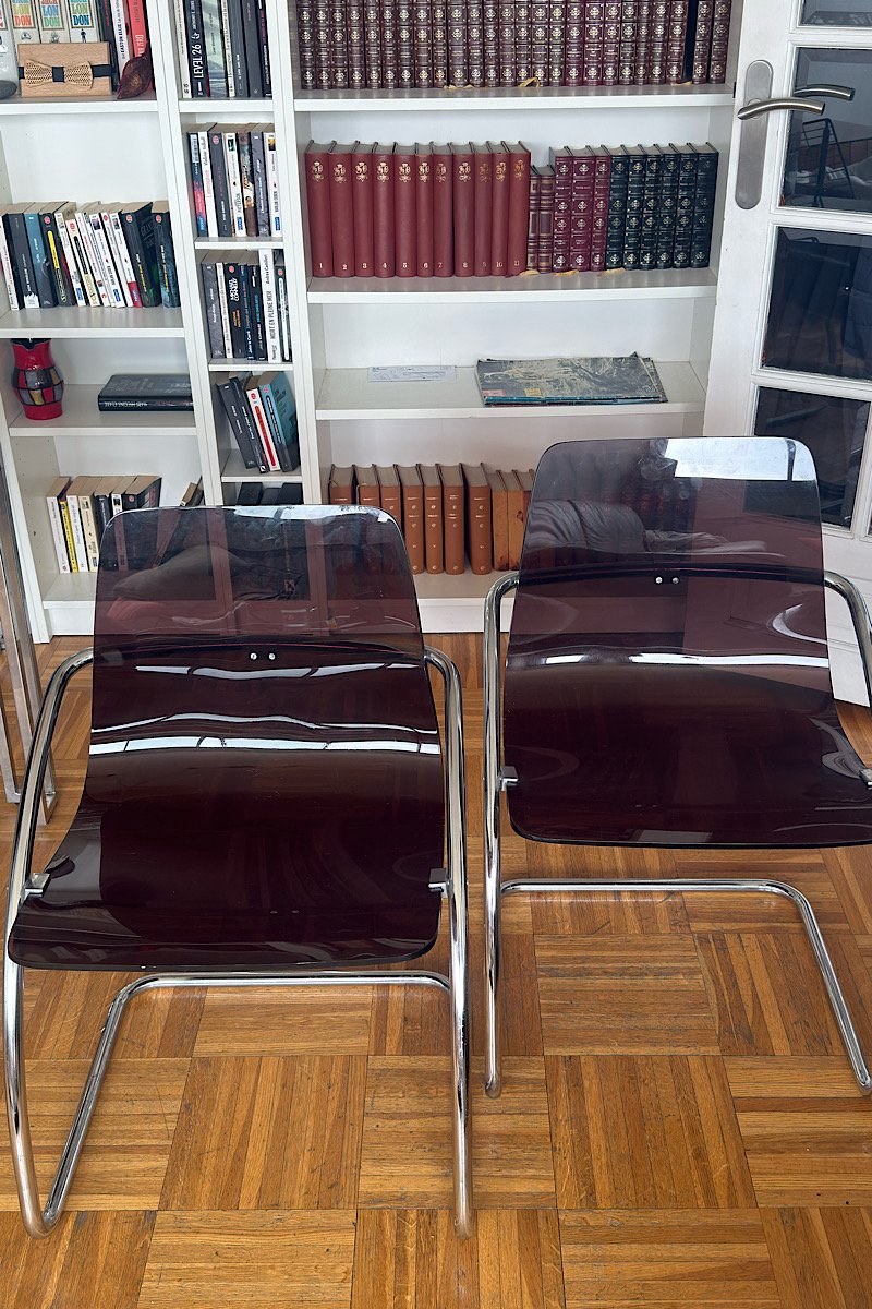 Deux chaises design "Blooming" créées par Yves Christin, dotées d'une structure métallique et d'une assise en plexiglas fumé, photographiées dans un intérieur avec parquet.