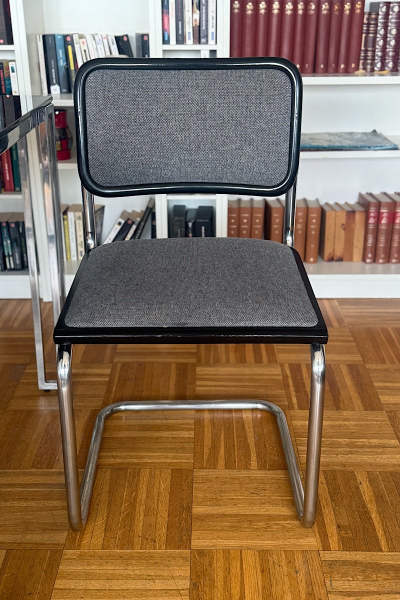 Vue de face d'une chaise Cesca B32 de Marcel Breuer avec son assise et son dossier en tissu gris, posée sur un sol en parquet et devant une bibliothèque