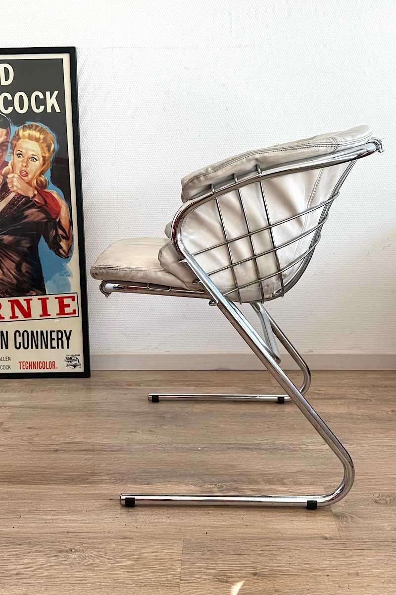 Vue de dos du fauteuil Flynn de Gastone Rinaldi, structure filaire en métal chromé et assise en cuir crème, dans un intérieur avec une affiche de film